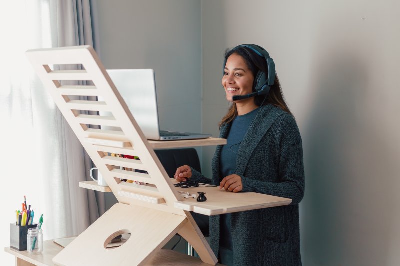 Young woman with long black hair and wearing black headphones working at a wooden standing desk facing a laptop in her home office (Photo by Justin - stock.adobe.com)